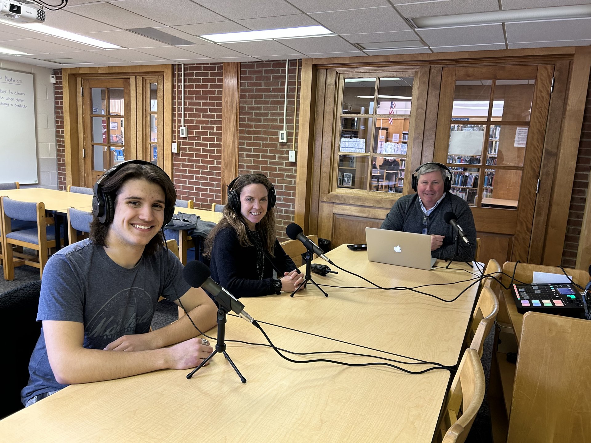 Elliott Lempke, Mrs. Bernard, and Mr. Konstant are seated at a table with headphones and microphones for podcasting.