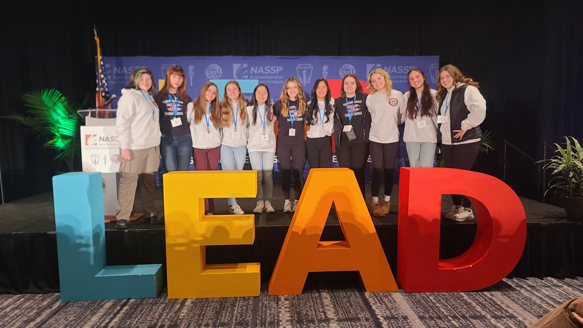 A group of students poses on stage behind large lettering spelling L E A D.