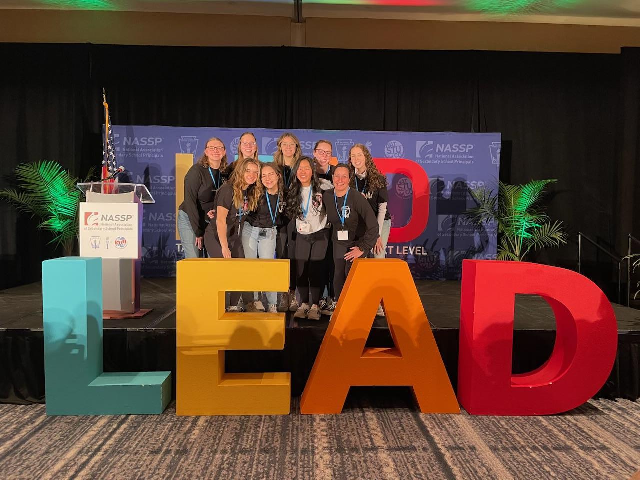 A group of students poses on stage behind large lettering spelling L E A D.